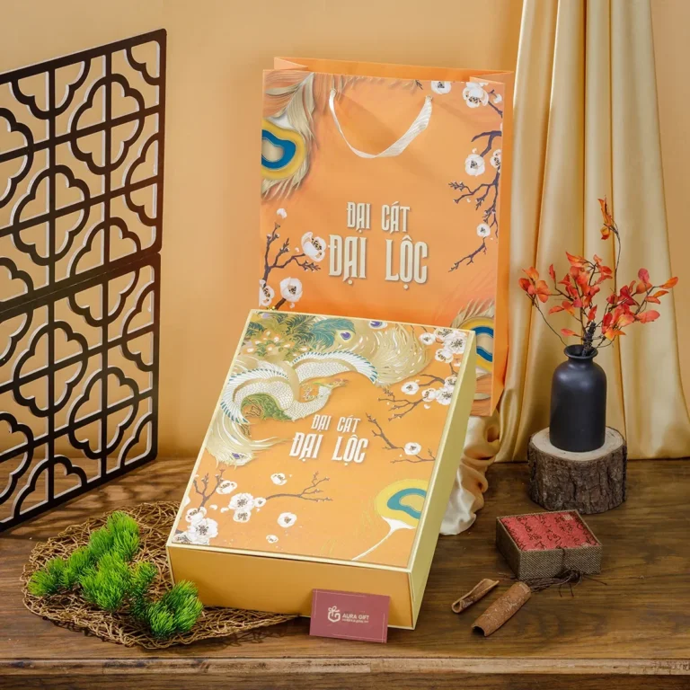 Hộp quà Tết màu cam in chim phượng và hoa mai đặt cạnh túi giấy cùng mẫu