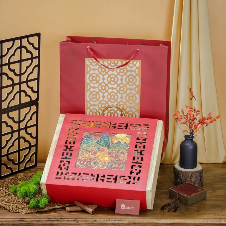 Hộp quà Tết mặt đỏ cắt rỗng song cửa, ô giữa tranh sơn thủy, đặt cạnh túi giấy cùng bộ